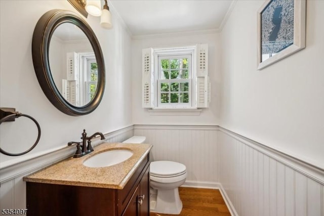 half bath featuring wood finished floors, a wainscoted wall, vanity, toilet, and ornamental molding