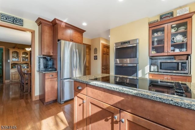 kitchen featuring brown cabinets, light wood finished floors, stone countertops, stainless steel appliances, and glass insert cabinets