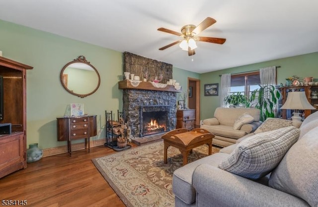 living area with baseboards, a stone fireplace, a ceiling fan, and wood finished floors