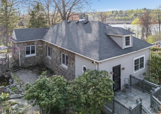 back of property with a shingled roof, a wooden deck, stone siding, and a chimney