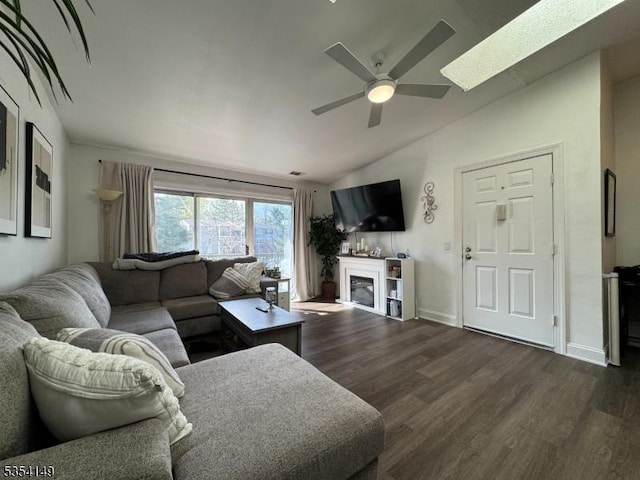 living area with a glass covered fireplace, a ceiling fan, baseboards, dark wood-type flooring, and vaulted ceiling