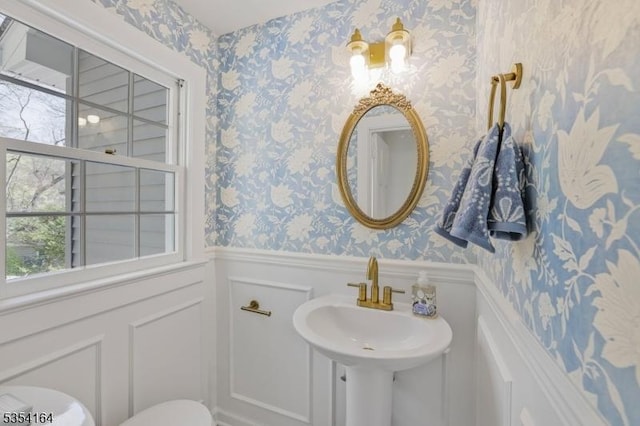 half bathroom featuring wainscoting, a sink, toilet, a decorative wall, and wallpapered walls