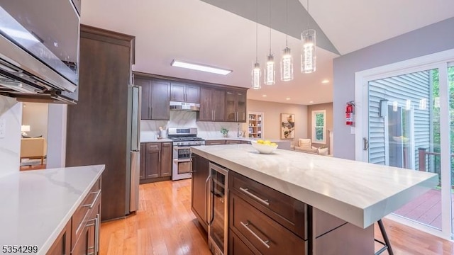 kitchen featuring stainless steel appliances, wine cooler, under cabinet range hood, light wood-style flooring, and hanging light fixtures
