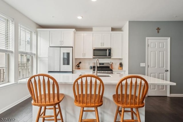kitchen featuring a kitchen island with sink, dark wood-style floors, light countertops, backsplash, and stainless steel appliances