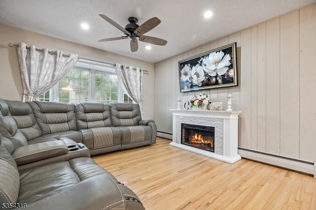 living area featuring baseboard heating, a glass covered fireplace, a ceiling fan, wood finished floors, and a baseboard radiator