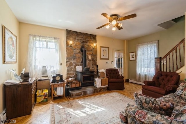 living room featuring stairs, a baseboard heating unit, a wood stove, ceiling fan, and baseboard heating