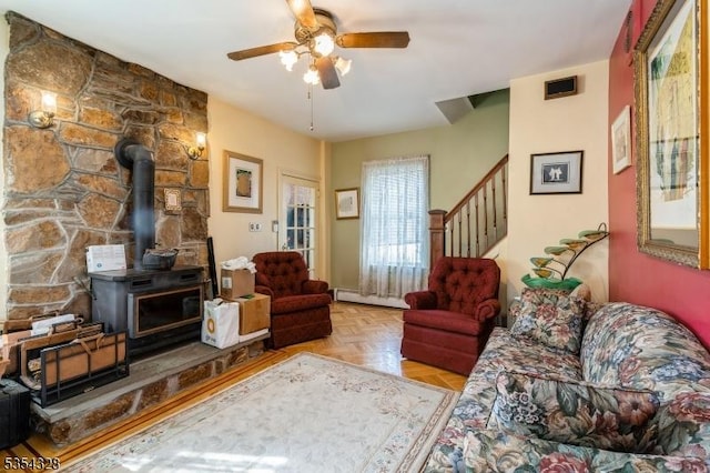 living room featuring a wood stove, baseboard heating, ceiling fan, and stairway