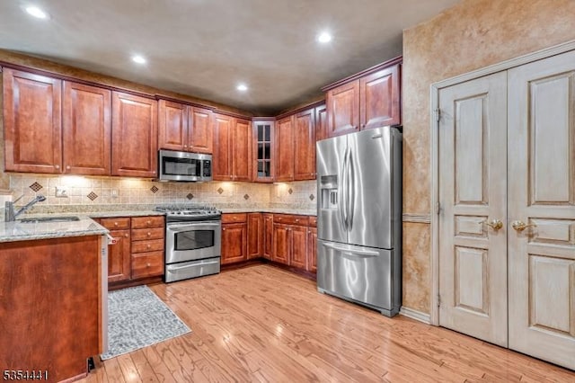kitchen featuring stainless steel appliances, a sink, light wood finished floors, light stone counters, and glass insert cabinets