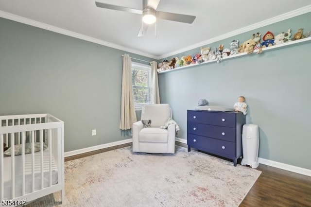 bedroom featuring crown molding, a nursery area, a ceiling fan, baseboards, and wood finished floors