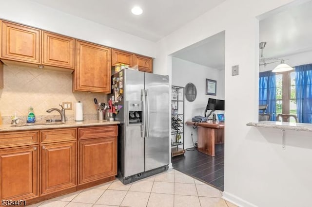 kitchen featuring brown cabinetry, backsplash, light stone countertops, a sink, and stainless steel fridge