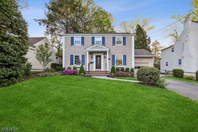 colonial house featuring an attached garage, aphalt driveway, a front yard, and a chimney