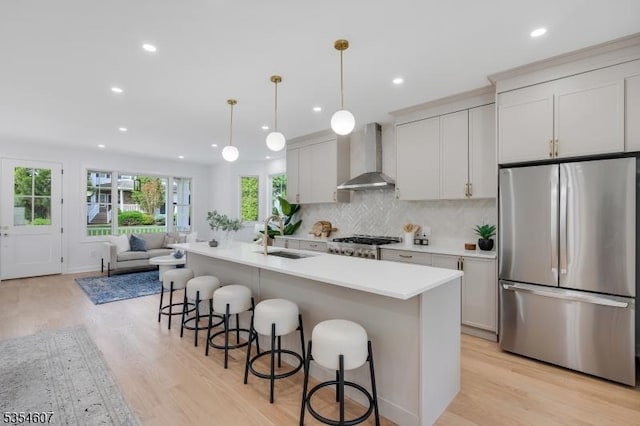 kitchen featuring stainless steel appliances, a sink, wall chimney exhaust hood, light wood-style flooring, and a kitchen bar