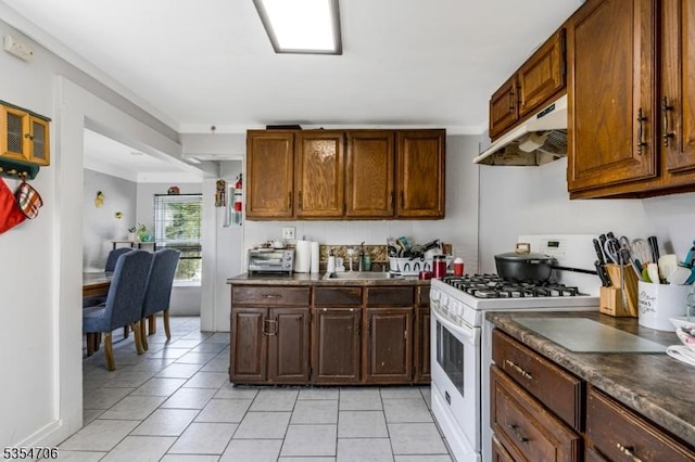 kitchen featuring under cabinet range hood, white range with gas stovetop, dark countertops, light tile patterned floors, and a sink