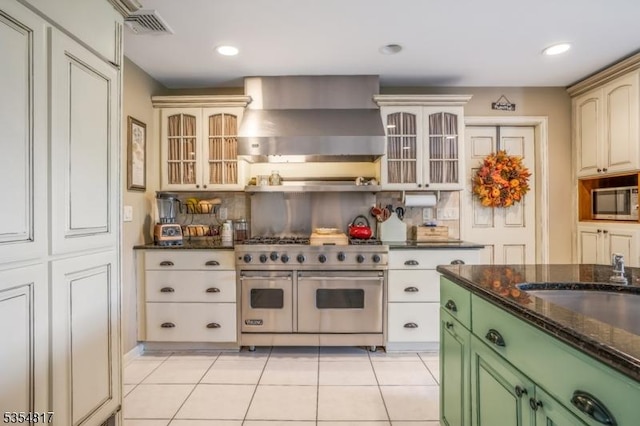 kitchen featuring wall chimney range hood, appliances with stainless steel finishes, light tile patterned floors, and glass insert cabinets