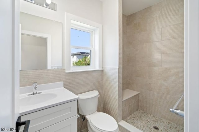 bathroom with a tile shower, vanity, toilet, a wainscoted wall, and tile walls