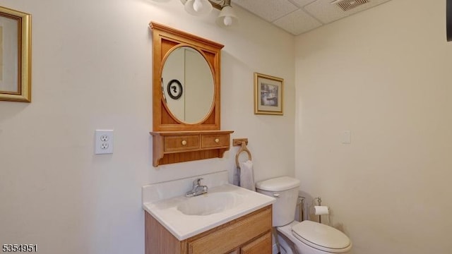half bathroom featuring visible vents, a drop ceiling, toilet, and vanity