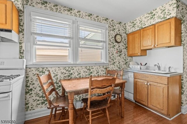 kitchen with white appliances, wallpapered walls, under cabinet range hood, wood finished floors, and a sink