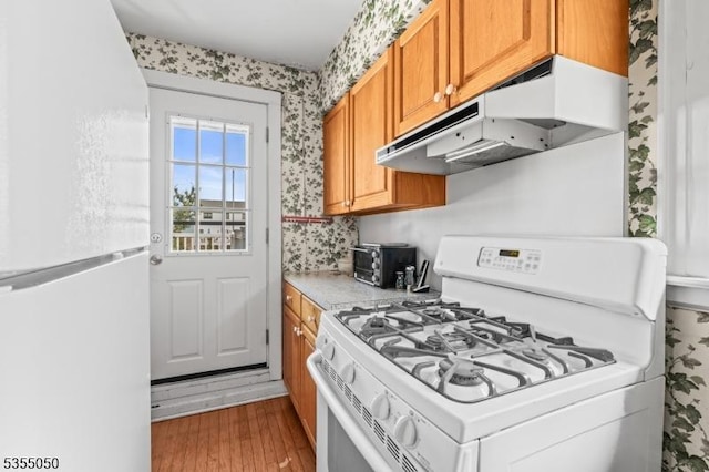 kitchen featuring white appliances, under cabinet range hood, wallpapered walls, wood finished floors, and brown cabinets