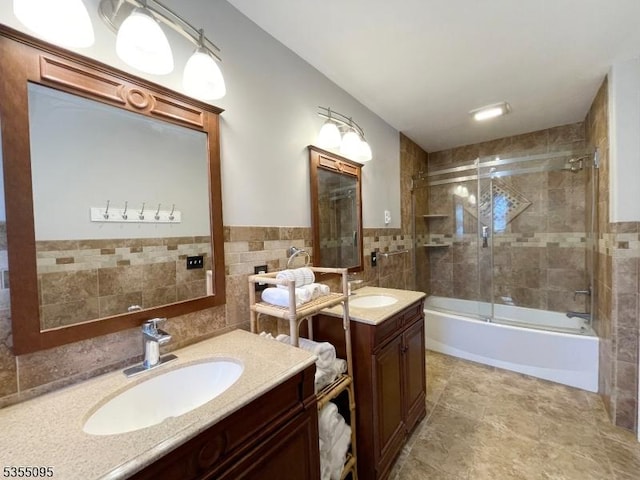 full bath with tile walls, a wainscoted wall, two vanities, a sink, and combined bath / shower with glass door