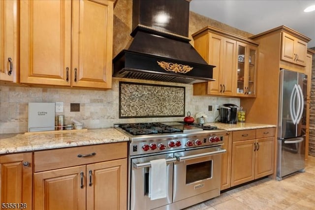 kitchen with stainless steel appliances, custom range hood, light stone counters, glass insert cabinets, and backsplash