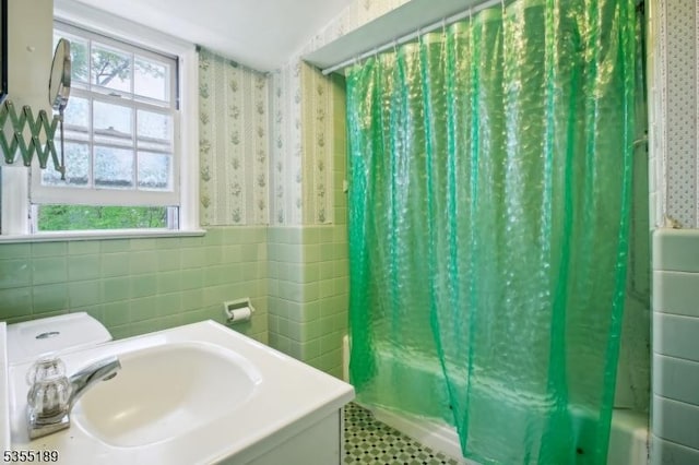 full bath with wallpapered walls, tile walls, a sink, wainscoting, and shower / bath combo