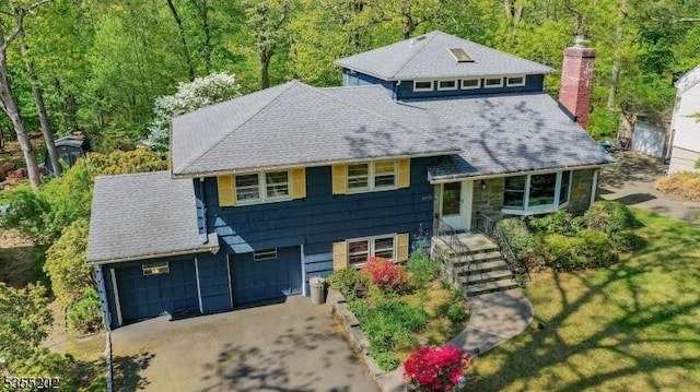 split level home with driveway, a chimney, roof with shingles, a view of trees, and an attached garage