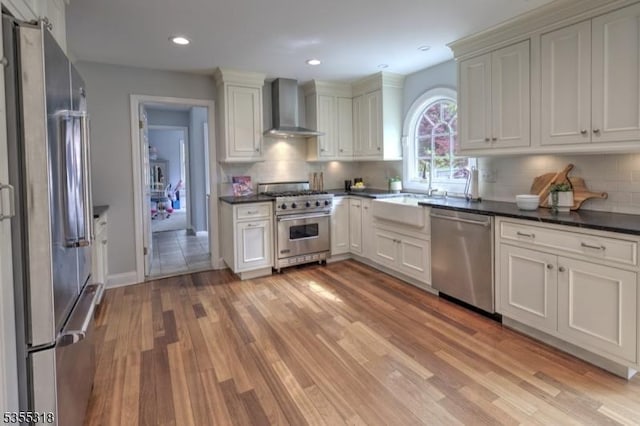 kitchen with wall chimney range hood, dark countertops, a sink, stainless steel appliances, and light wood-type flooring