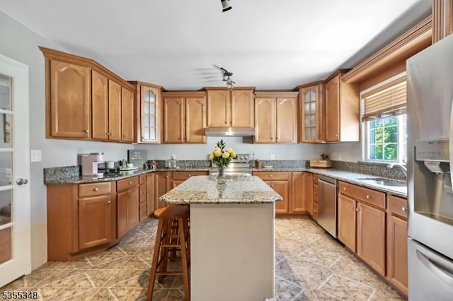 kitchen with appliances with stainless steel finishes, under cabinet range hood, a center island, light stone counters, and glass insert cabinets