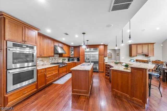 kitchen featuring a kitchen island with sink, brown cabinetry, stainless steel appliances, under cabinet range hood, and visible vents
