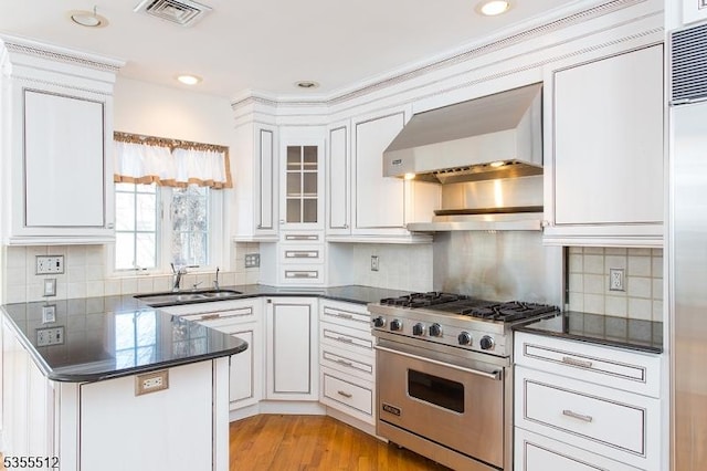 kitchen with high quality appliances, wall chimney exhaust hood, dark countertops, a sink, and white cabinets