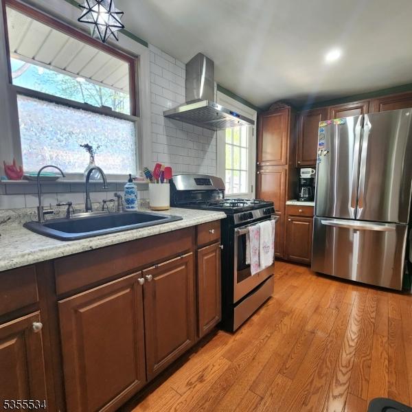 kitchen with wall chimney range hood, tasteful backsplash, a sink, light wood finished floors, and stainless steel appliances