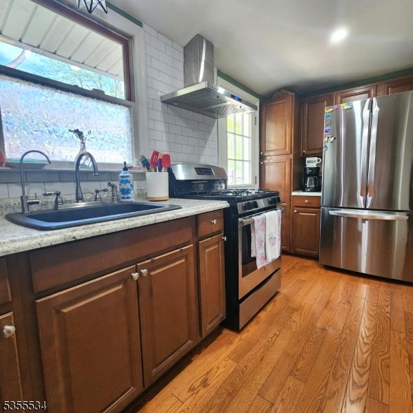 kitchen with a sink, light countertops, light wood-style flooring, wall chimney exhaust hood, and stainless steel appliances