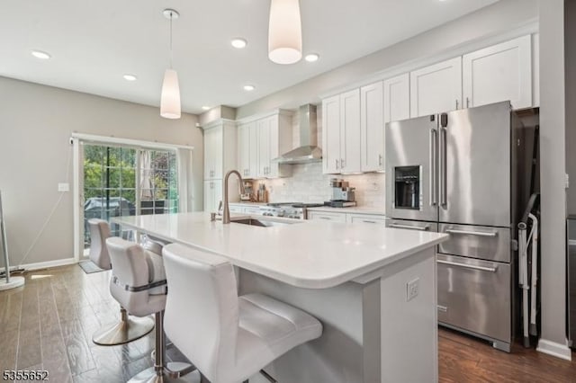 kitchen with high end fridge, baseboards, a sink, wall chimney range hood, and a center island with sink