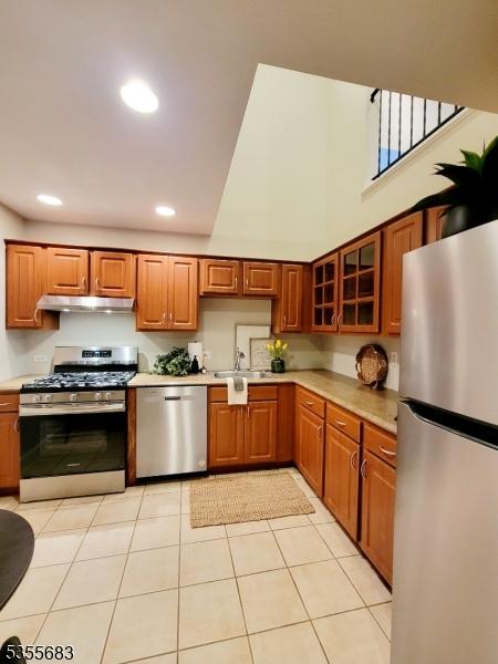 kitchen with appliances with stainless steel finishes, under cabinet range hood, light countertops, brown cabinetry, and a towering ceiling