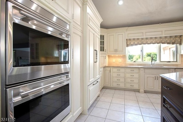 kitchen with a sink, double oven, light tile patterned flooring, light stone counters, and backsplash