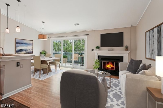living area with lofted ceiling, light wood-style flooring, ornamental molding, a glass covered fireplace, and visible vents