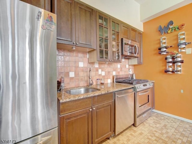 kitchen featuring light stone countertops, glass insert cabinets, backsplash, brown cabinets, and appliances with stainless steel finishes