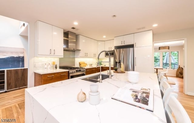 kitchen featuring appliances with stainless steel finishes, wall chimney exhaust hood, a sink, white cabinets, and light wood-style flooring
