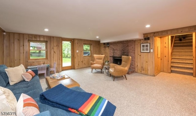 living room featuring wooden walls, stairway, a brick fireplace, carpet flooring, and healthy amount of natural light
