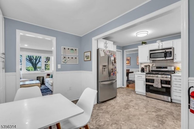 kitchen with stainless steel appliances, white cabinetry, wainscoting, light countertops, and crown molding