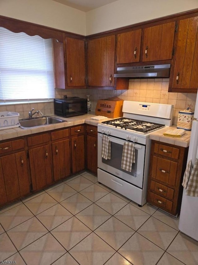 kitchen featuring white gas stove, black microwave, under cabinet range hood, tile countertops, and a sink