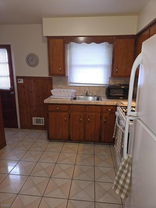 kitchen featuring a sink, white appliances, light countertops, a wainscoted wall, and brown cabinetry