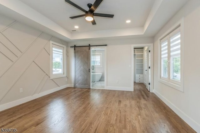 unfurnished bedroom featuring a raised ceiling, wood finished floors, a barn door, baseboards, and a walk in closet