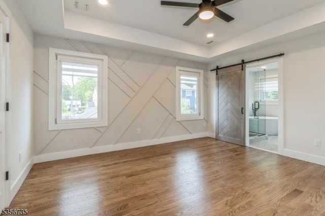 unfurnished bedroom with a barn door, baseboards, a raised ceiling, wood finished floors, and connected bathroom