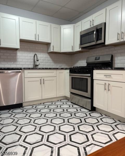 kitchen featuring appliances with stainless steel finishes, a sink, dark countertops, decorative backsplash, and a drop ceiling