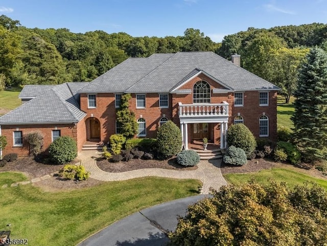 colonial-style house with brick siding, a chimney, a front lawn, a balcony, and a view of trees