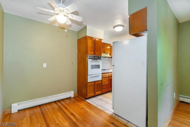 kitchen featuring white appliances, a baseboard radiator, a warming drawer, under cabinet range hood, and brown cabinetry
