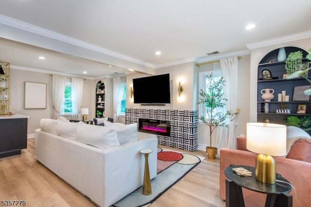 living area with light wood-style floors, recessed lighting, a glass covered fireplace, crown molding, and built in shelves