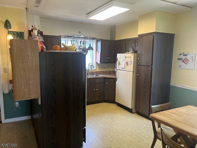 kitchen with freestanding refrigerator, dark brown cabinetry, a sink, crown molding, and baseboards