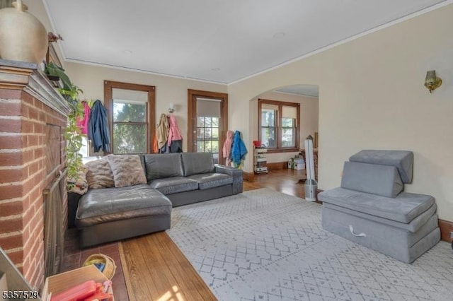 living room with arched walkways, a brick fireplace, plenty of natural light, wood finished floors, and ornamental molding